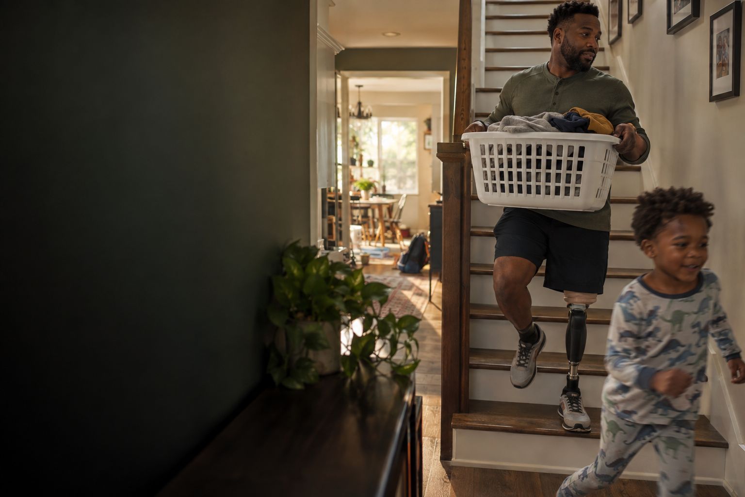 A father with an above-knee prosthesis carrying a laundry basket up the stairs as his son runs past (concept image)